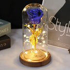 A preserved glittery blue rose with glowing gold fairy lights inside a clear glass dome, displayed on a round wooden base.